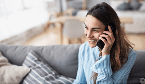 Frau mit braunen Haaren sitzt auf grauem Sofa und hält ein Telefon ans Ohr